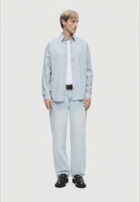 Ανοιχτό μπλε ριγέ πουκάμισο με κουμπιά πάνω από ένα λευκό μπλουζάκι, σε συνδυασμό με ανοιχτό μπλε τζιν και μαύρα παπούτσια, στέκεται σε ουδέτερο φόντο.