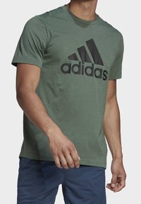 Homme portant un t-shirt Adidas vert avec un grand logo noir et un short bleu marine, coupé au niveau du cou et des cuisses.