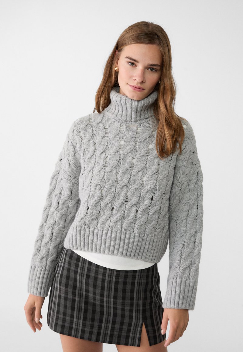 Maglione grigio a maglia con colletto alto e maniche lunghe, abbinato a una mini gonna a quadri nera con spacco laterale.