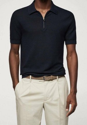 Mężczyzna ubrany w czarną, krótkorękawową dzianinową koszulkę polo z frontowym zamkiem błyskawicznym, beżowe spodnie i jasnobrązowy pasek, ręce w kieszeniach.