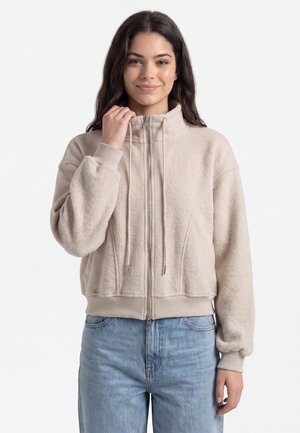 Giovane donna dai lunghi capelli scuri che indossa una giacca in pile beige con zip e jeans blu, tiene il colletto con una mano e sorride dolcemente.