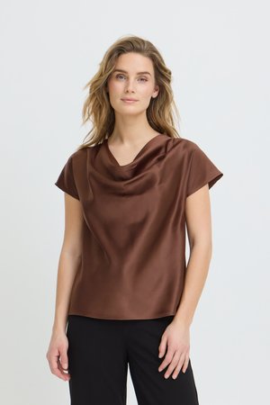 Sieviete ar viļņainiem blondiem matiem valkā brūnu satīna īsroku topu ar drapētu izgriezumu un melnas bikses, stāvot pret gaišu fonu.