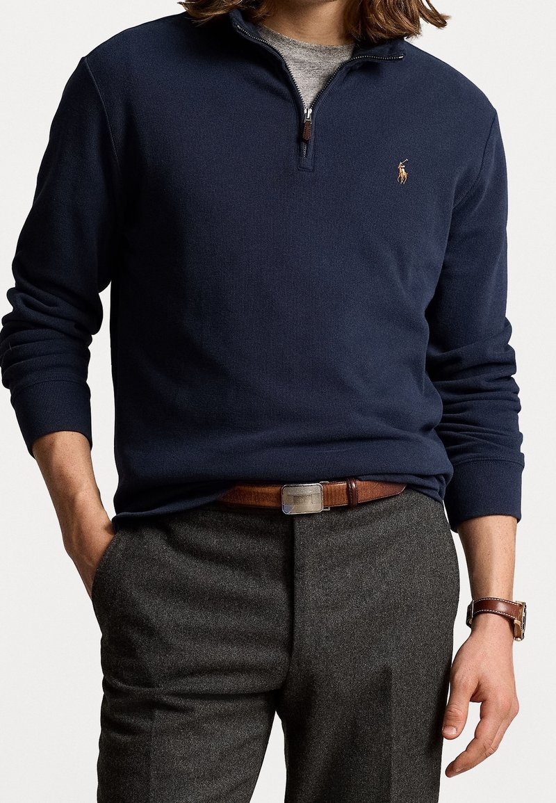 Mann in navyfarbener Quarter-Zip-Pullover, grauen Wollhosen, braunem Ledergürtel und braunem Uhrenarmband, mit der linken Hand in der Tasche.