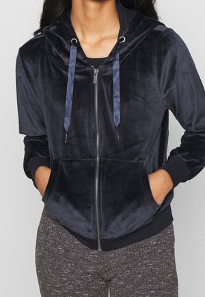 Nainen, joka käyttää tummansinistä veluurista vetoketjullista hupparia satiininauhoilla ja harmaita teksturoituja housuja, kädet hupun taskuissa.