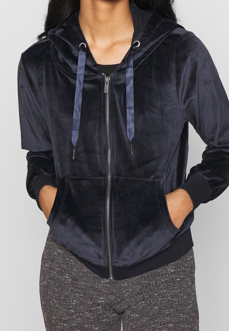 Femme portant un sweat à capuche zippé en velours bleu marine foncé avec des cordons de serrage en satin et un pantalon texturé gris, les mains dans les poches du sweat à capuche.