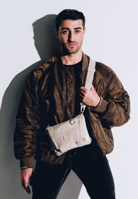 Borsa a tracolla beige con un design strutturato, dotata di una tasca frontale e spallacci regolabili, indossata sopra una giacca bomber marrone.