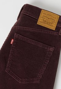 Σκούρο καφέ βελούδινο παντελόνι Levi's διπλωμένο ώστε να φαίνεται η πίσω τσέπη, η κόκκινη ετικέτα Levi's και η ταμπέλα από ταμπά δέρμα της μάρκας Levi Strauss.