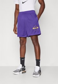 Lilla võrgustikuga korvpallishorts, millel on kollane "LAKERS" logo, elastne vöökoht ja küljetaskud, kombineerituna mustade spordisokkide ja -kingadega.