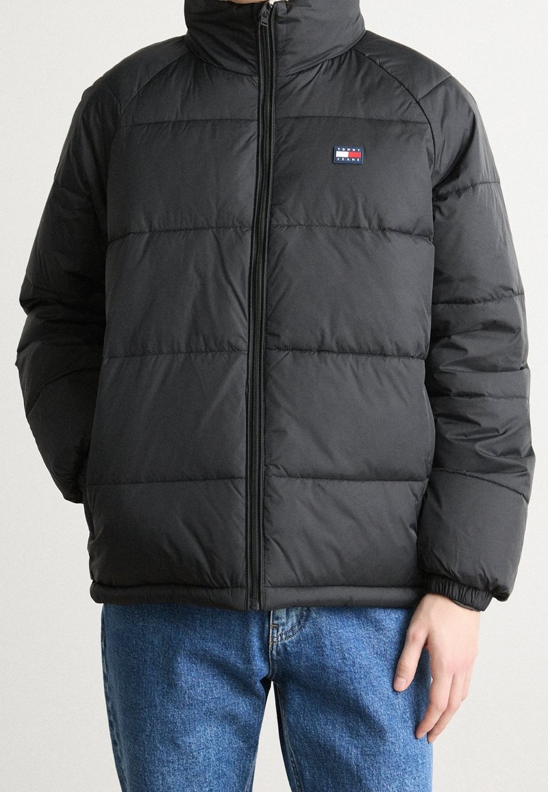 Homme portant une veste matelassée noire zippée avec le logo Tommy Jeans sur la poitrine et un jean bleu, debout devant un fond clair.