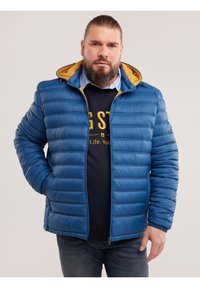 Szakállas férfi hátrafésült hajjal, kék puffert visel sárga kapucni béléssel és sötét farmerben, egyik kezét a zsebében tartva áll.