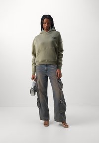 Felpa verde oliva con tasca anteriore e testo nero; pantaloni cargo blu chiaro con tasche laterali; sandali con tacco trasparente; borsa argentata.