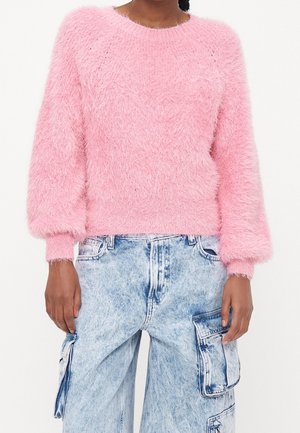 Personne portant un pull duveteux rose clair avec des manches ballon et un jean cargo bleu clair délavé à l'acide, debout devant un fond blanc uni.
