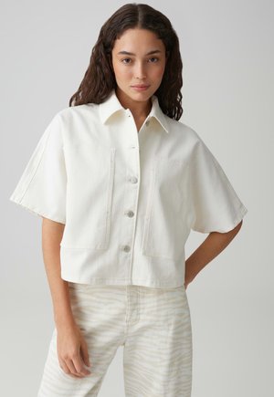 Junge Frau mit langen lockigen Haaren, die ein weißes, bauchfreies Hemd mit Knöpfen und helle, gemusterte Hosen vor einem einfarbigen Hintergrund trägt.