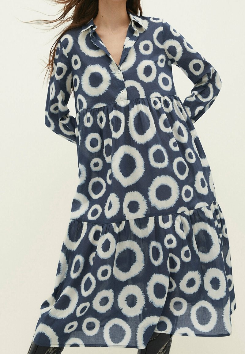 Robe bleu marine avec des motifs circulaires blancs irréguliers, manches longues, jupe à volants et col partiellement ouvert.
