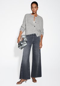 Cardigan grigio lavorato a maglia con scollo rotondo e bottoni neri, abbinato a jeans scuri a gamba larga. Accessoriato con una borsa metallica e sandali.