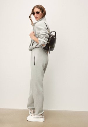Pantalon de survêtement en polaire grise et pull assorti à la coupe oversize. Mini sac à dos noir, baskets blanches avec un motif texturé et lunettes de soleil.