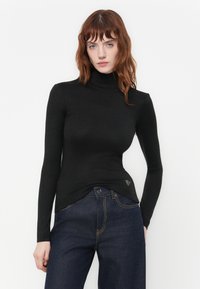 Donna con capelli castani mossi che indossa un maglione nero a collo alto e jeans blu scuro su uno sfondo bianco semplice.
