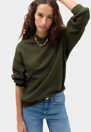 Kvinde med krøllet hår iført en olivengrøn sweatshirt, guldkæde halskæde og blå jeans, løfter en arm over hovedet.