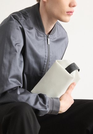 Gray bomber jacket with ribbed collar, holding a white silicone bag with a black strap. Smooth texture, minimalistic design.