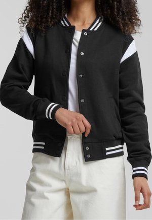 Personne portant une veste universitaire noire avec des rayures blanches sur le col, les poignets et les épaules, associée à un pantalon blanc et une chemise blanche.