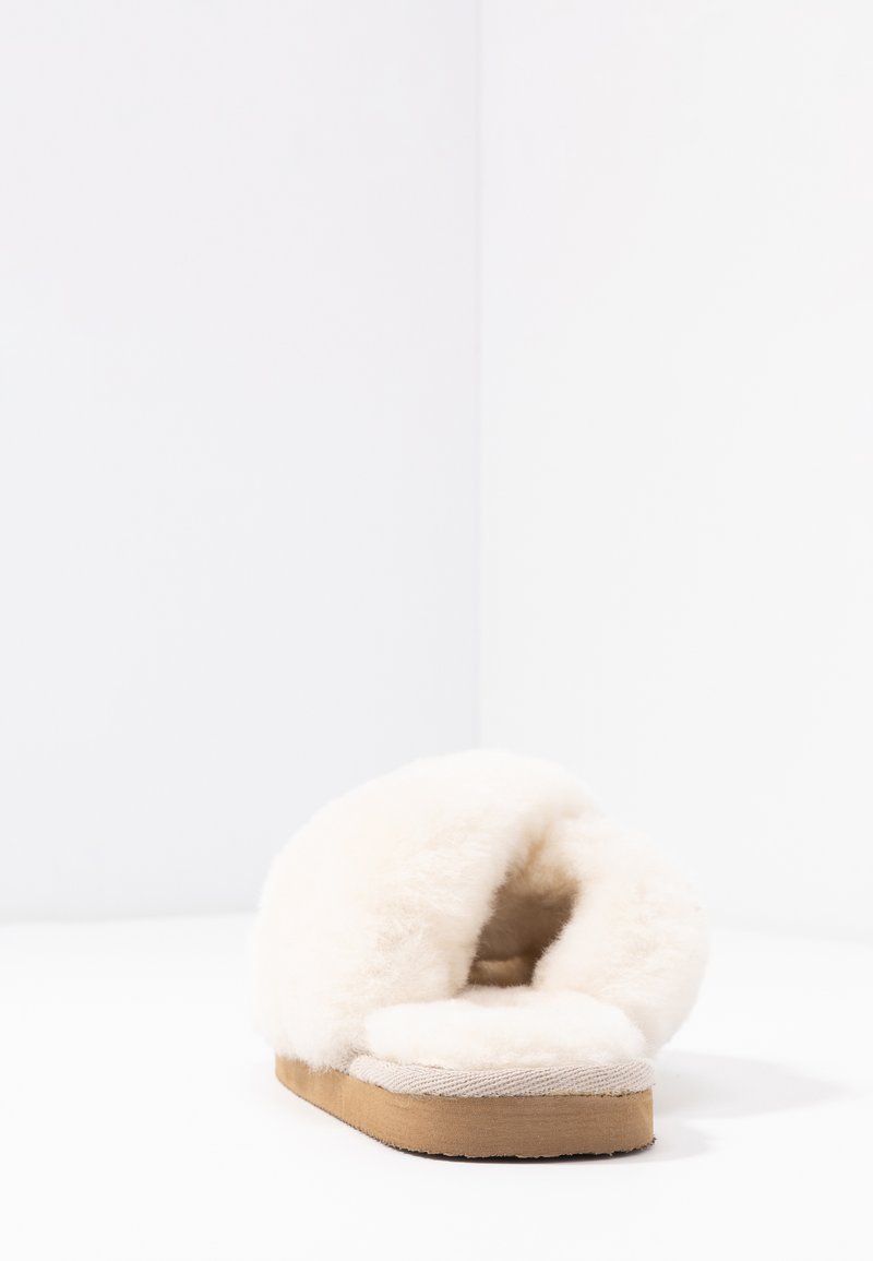 Crème-kleurige slipper met een pluizige bont bovenkant, open achterkant en een platte, licht tan kleurige zool. Zachte textuur, eenvoudig ontwerp en minimale details.