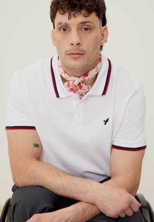 Άνδρας με κοντά κατσαρά μαλλιά που φοράει λευκή πόλο μπλούζα με κόκκινη και ναυτική διακόσμηση, φλοράλ μαντήλι στον λαιμό, κάθεται σε αναπηρικό καροτσάκι και κοιτάζει μπροστά.