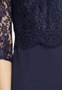 Námořnická krajková halenka s květinovými vzory na pevném námořnickém podkladu, ukazující detailní krajkové rukávy a zvlněný krajkový lem na oděvu.