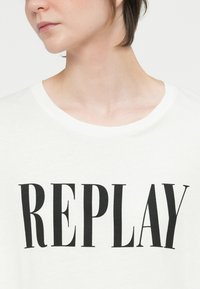 T-shirt de algodão branco com a palavra em negrito, preta, "REPLAY" impressa na parte da frente. Gola redonda e mangas curtas. Textura suave.