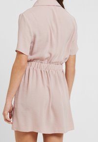 Donna che indossa un abito rosa chiaro a maniche corte con elastico in vita, mostrata di spalle contro uno sfondo neutro.