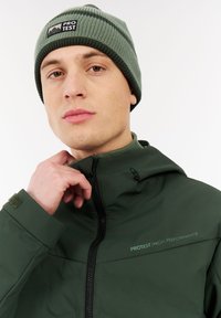 Žalias nepralaidus striukė su užtrauktuku priekyje, aukštu apykakle ir reguliuojama kapišonu; derinama su atitinkama žalia kepure, turinčia horizontalių juostų.