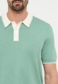 Άνδρας που φοράει κοντομάνικο πόλο μεντας πράσινου χρώματος με λευκό γιακά και λευκά κουμπιά, εμφανίζοντας το στήθος και την περιοχή του λαιμού.