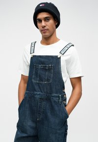 Denim-Latzhose mit einer vorderen Tasche, verstellbaren Trägern mit Logoakzenten und seitlichen Knöpfen. Getragen über einem schlichten weißen T-Shirt.
