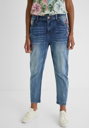 Kobieta ubrana w wyblakłe niebieskie krótkie dżinsy z kwiatowym haftem i białe sznurowane trampki, w połączeniu z kwiatowym bezrękawnikiem.