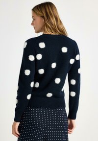 Maglione blu navy con grandi pois bianchi, caratterizzato da una finitura morbida e strutturata. Polsini e orlo a coste, indossato con una gonna a pois.