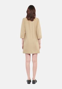 Khaki short dress with three-quarter sleeves, a straight cut, and a clean back seam. Worn with black footwear, against a plain background.