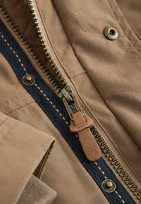 Tan fabric with a quilted design features a bronze zipper and suede pull tab, complemented by navy stitching and a metal snap button.