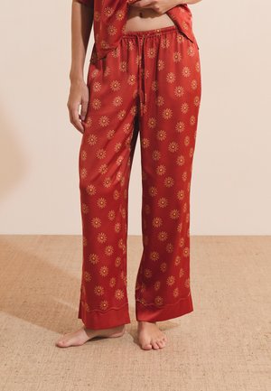 Persoon draagt losse rode satijnen broek met een geel bloemenpatroon, staat op blote voeten op een beige gestructureerde vloer.