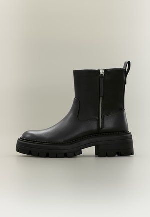 Bottine en cuir noir avec fermeture éclair latérale, semelle épaisse cramponnée, et languette à l’arrière, présentée sur un fond neutre.