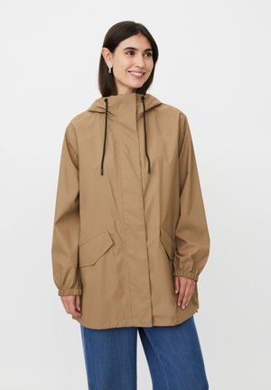 Kobieta ubrana w beżową kurtkę przeciwdeszczową z kapturem i czarnymi troczkami oraz niebieskie dżinsy, stojąca na tle białego, jednolitego tła.