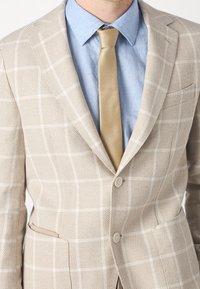Beżowa marynarka w kratę z teksturowanej tkaniny, z dwoma guzikami i subtelnymi klapami, w zestawieniu z jasnoniebieską koszulą i kremowym krawatem.