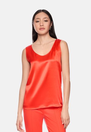 Woman with long dark hair wearing shiny red sleeveless top and matching red pants, standing against a plain light background.
