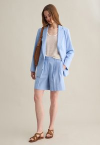 Blazer su misura di un azzurro chiaro con pantaloni shorts abbinati, top strutturato di un bianco sporco, borsa a tracolla marrone e sandali marroni con cinturini.