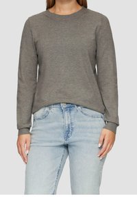 Kvinna som bär en enkel grå långärmad tröja och ljusblå högmidjade jeans, stående mot en neutral bakgrund.