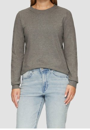 Femme portant un pull gris uni à manches longues et un jean taille haute bleu clair, se tenant devant un fond neutre.