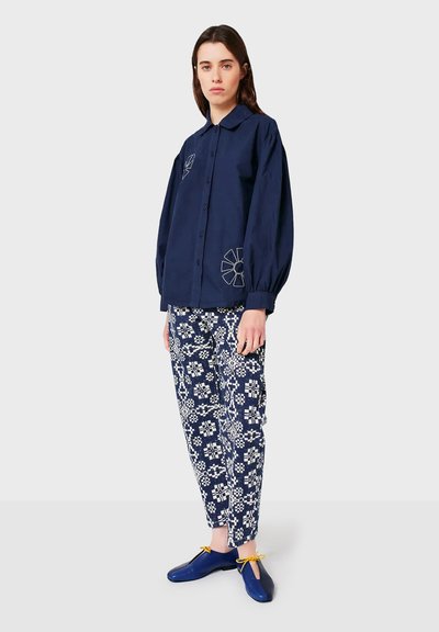 Donna che indossa una camicia blu scuro con bottoni e design di fiori ricamati, pantaloni a fantasia blu e bianchi, e scarpe blu con lacci gialli, in piedi.