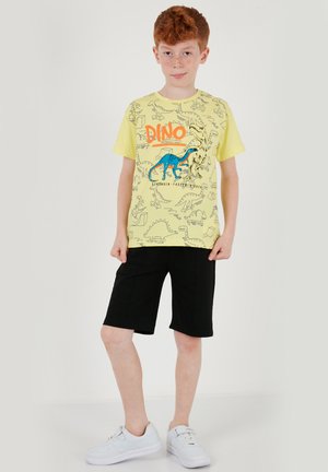 Un niño pelirrojo está de pie, vistiendo una camiseta amarilla con estampado de dinosaurios, pantalones cortos negros y zapatillas blancas, sobre un fondo liso.