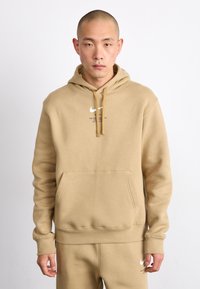 Sweatshirt Nike bege com um bolso frontal e cordão. Apresenta um logotipo e texto branco no peito. Feito de um tecido macio e texturizado.
