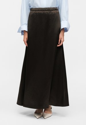 Kobieta ubrana w ciemnobrązową spódnicę o wysokim stanie, sięgającą do kostek, z jasno niebieską koszulą z długim rękawem i beżowymi, szpiczastymi butami.