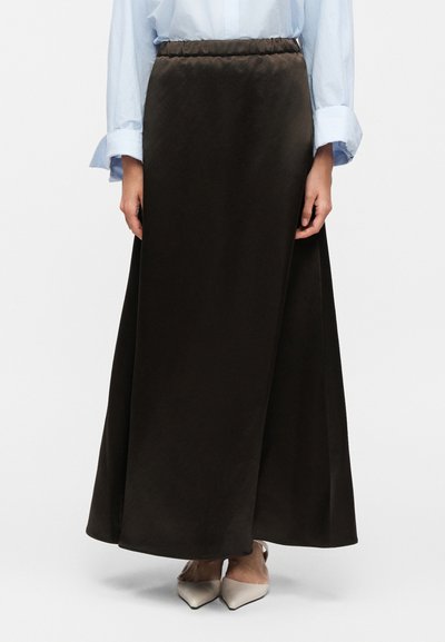 Femme portant une jupe marron foncé taille haute, longueur cheville, avec une chemise bleu clair à manches longues et des chaussures beiges pointues.