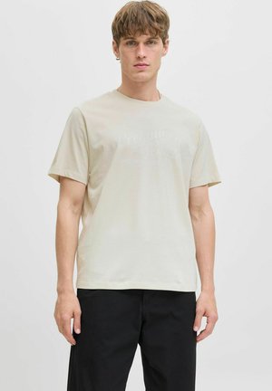 Cremfarbendes Baumwoll-T-Shirt mit Rundhalsausschnitt und kurzen Ärmeln. Mit einem dezenten grafischen Druck auf der Vorderseite. Kombiniert mit schwarzen Hosen.